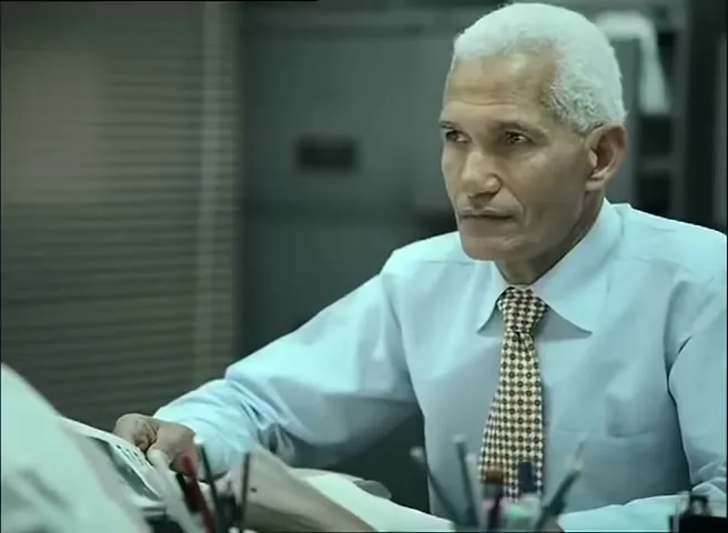 An older man with white hair and a light blue shirt sits at a desk, looking to the side in an office.