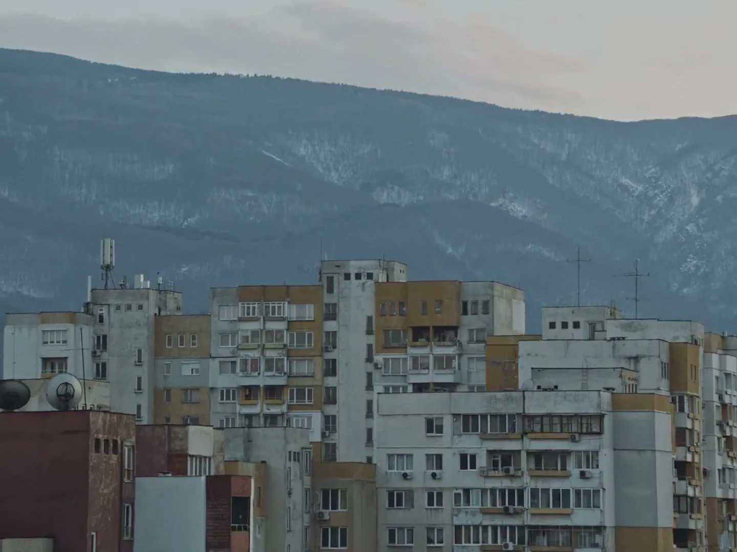 A wide shot of a city with old, worn-down apartment buildings in the foreground and a large, snow-capped mountain in the background under a cloudy sky.