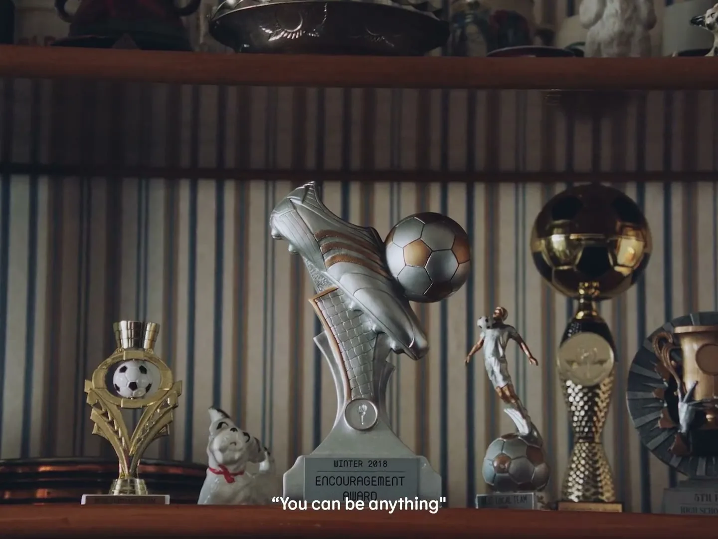 A collection of soccer trophies and awards are displayed on a wooden shelf against a striped wallpaper background, with one trophy prominently featuring a soccer boot and ball.