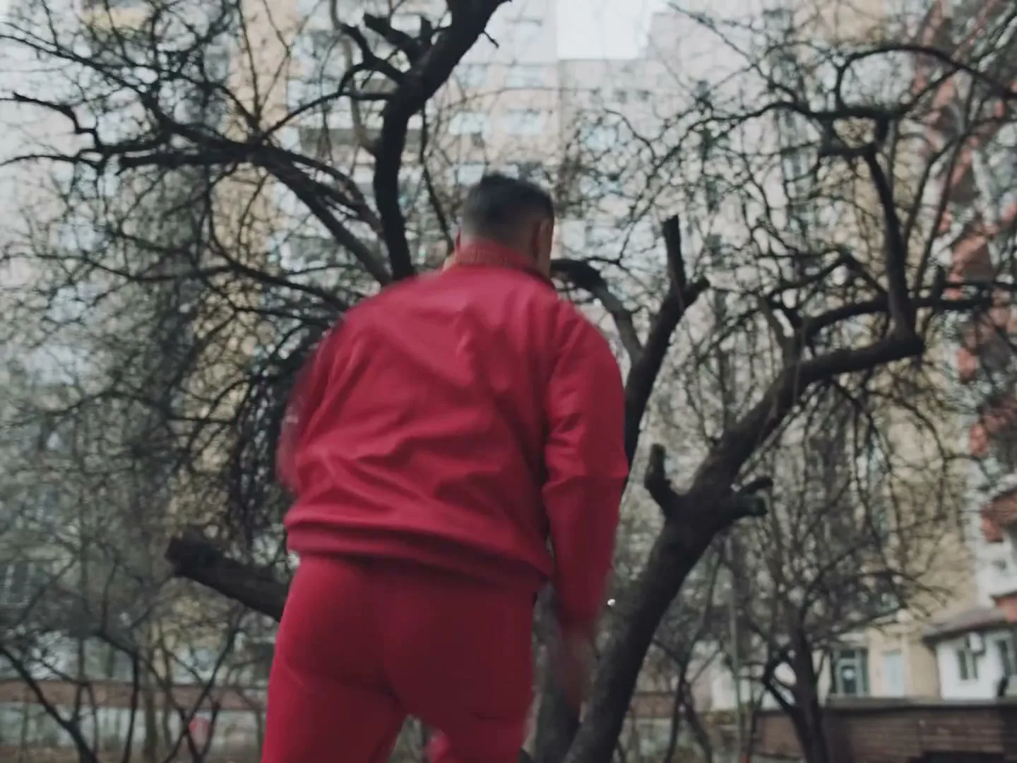 A man in a red tracksuit runs and jumps over a low wall in an urban park, with bare trees and apartment buildings in the background.