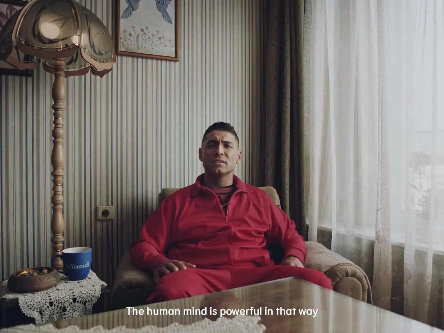 A man in his 30s, wearing a red tracksuit, sits in an armchair in a dimly lit living room, looking directly at the camera and speaking.