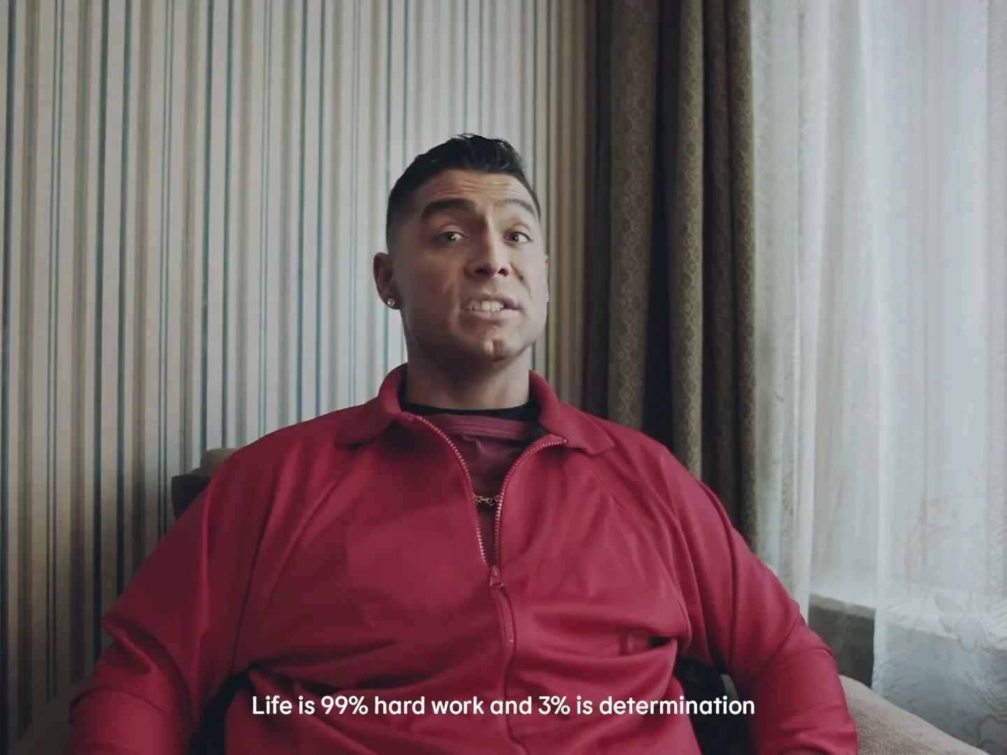 A man in his 30s, wearing a red tracksuit, sits in a chair and speaks directly to the camera in an interior room.