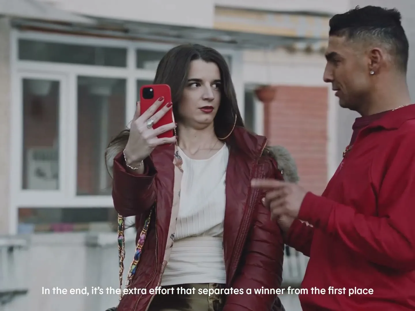 A young woman in a red puffer jacket takes a selfie with a red phone while a young man in a red hoodie talks to her on a residential street.