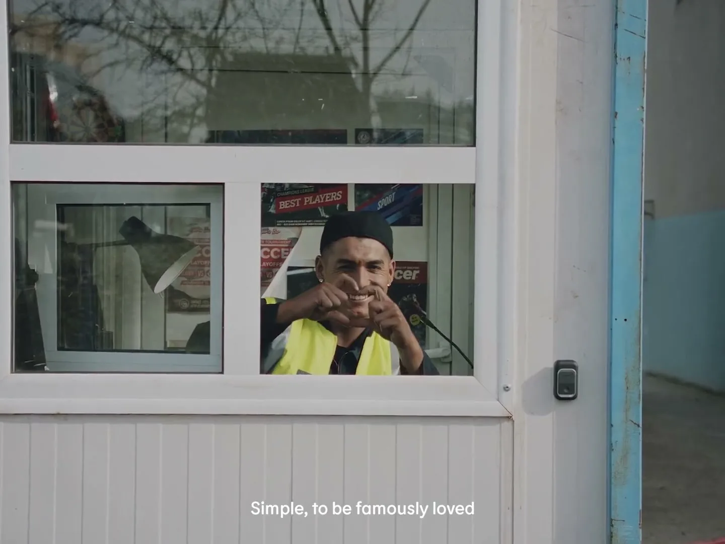 A man in a black beanie and yellow vest smiles and makes a heart shape with his hands through a window of a small building.