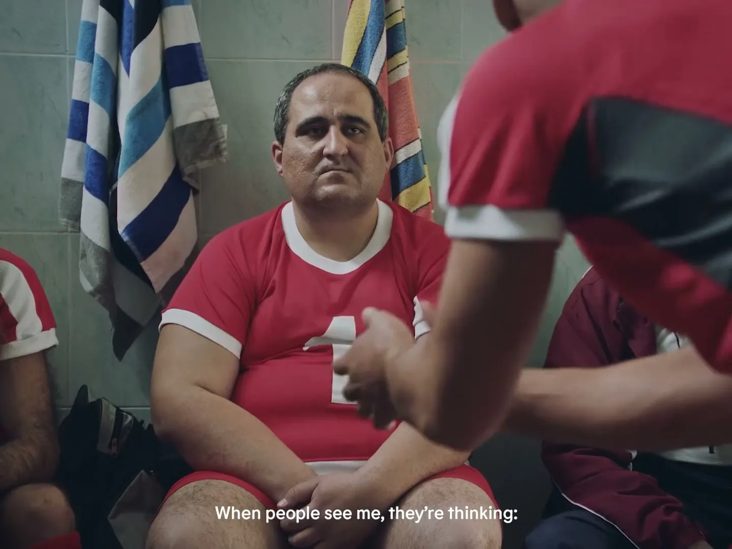 A man in a red soccer jersey with the number 1 sits on a bench in a locker room, looking serious, while another man's arm is blurred in the foreground.