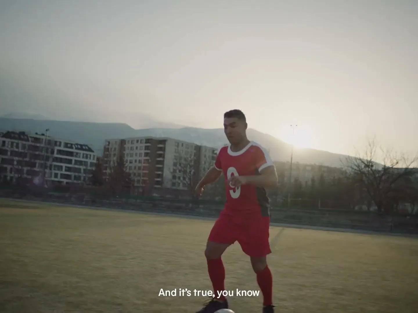 A male soccer player in a red uniform dribbles a soccer ball on a field with buildings and mountains in the background at sunset.