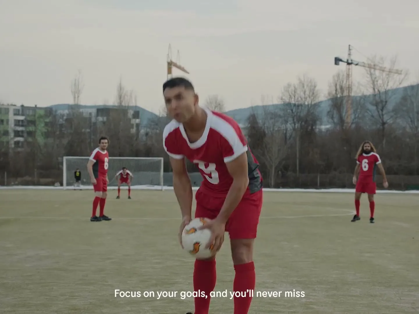 A male soccer player in a red uniform places a soccer ball on the field, surrounded by teammates, on a cloudy day.