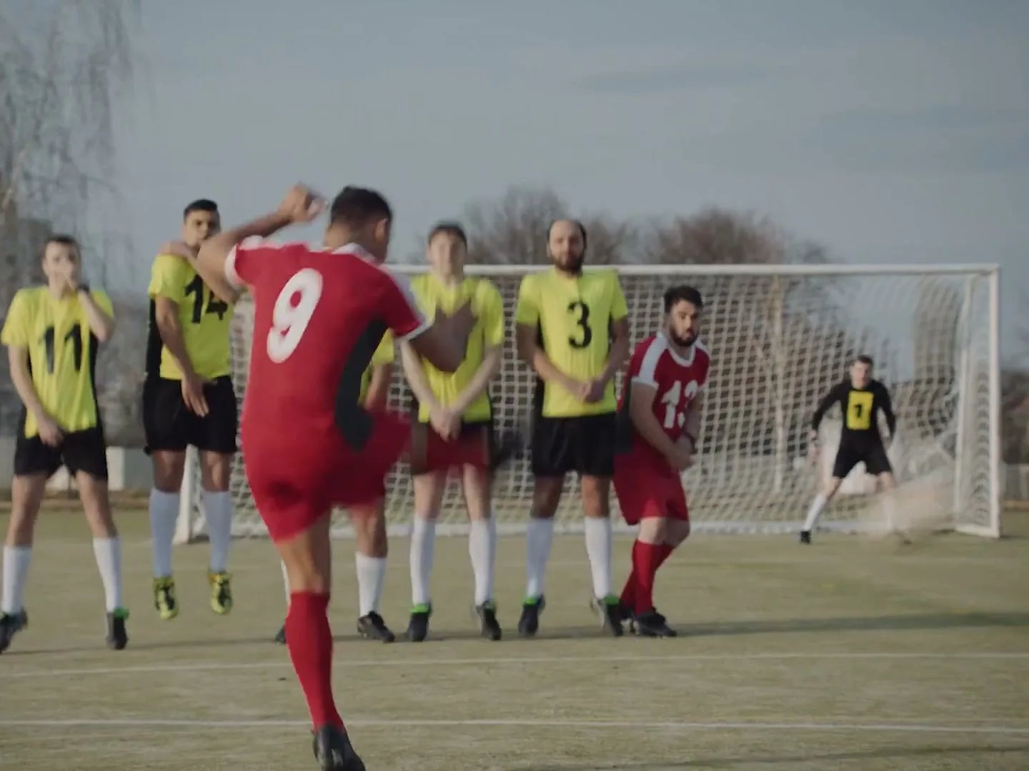 A male soccer player in a red jersey kicks a soccer ball towards a goal, while other players in yellow and red jerseys stand in a line.