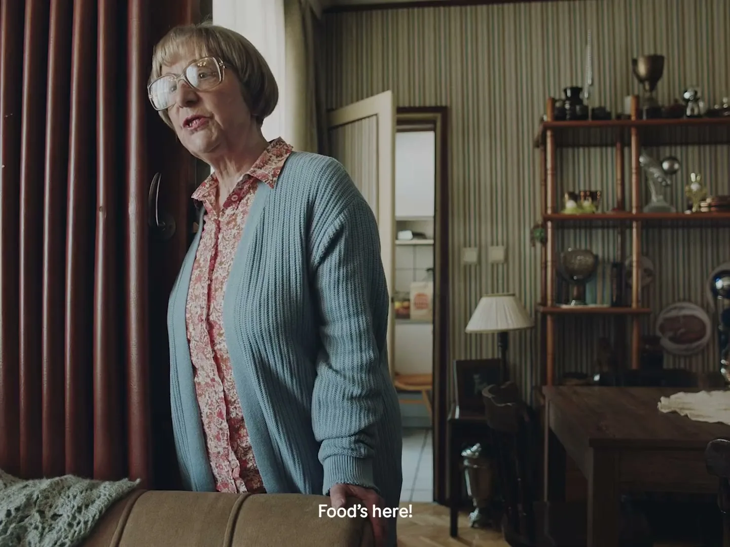 An elderly woman with glasses and a blue cardigan looks out a window in a cozy, old-fashioned living room.