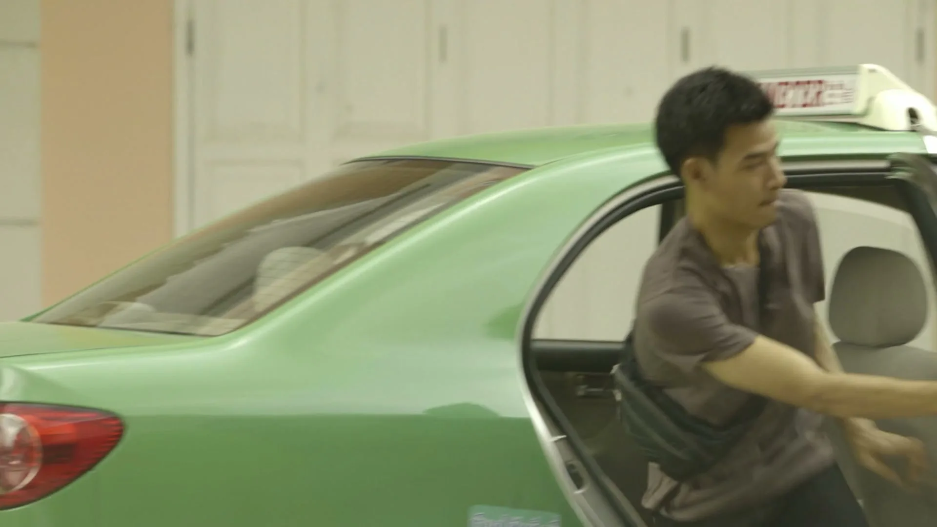 A young man in a brown t-shirt and dark pants exits a green taxi cab on a residential street during the day.