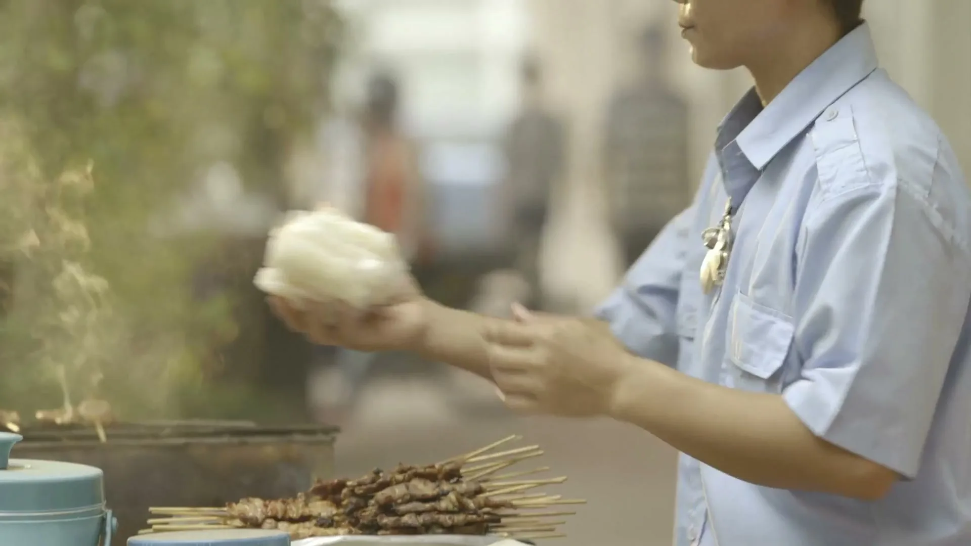 A male street vendor in a light blue shirt prepares and hands over food to a customer on a busy street.