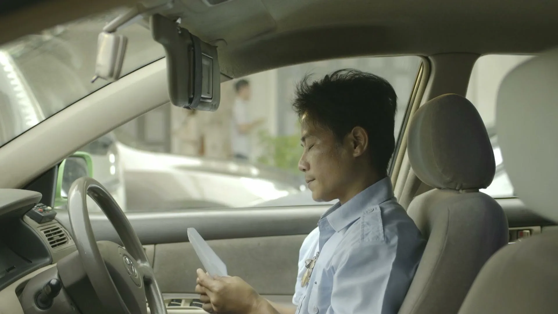 A male taxi driver in a light blue shirt sits in his car, looking at a piece of paper, then puts it in the sun visor.
