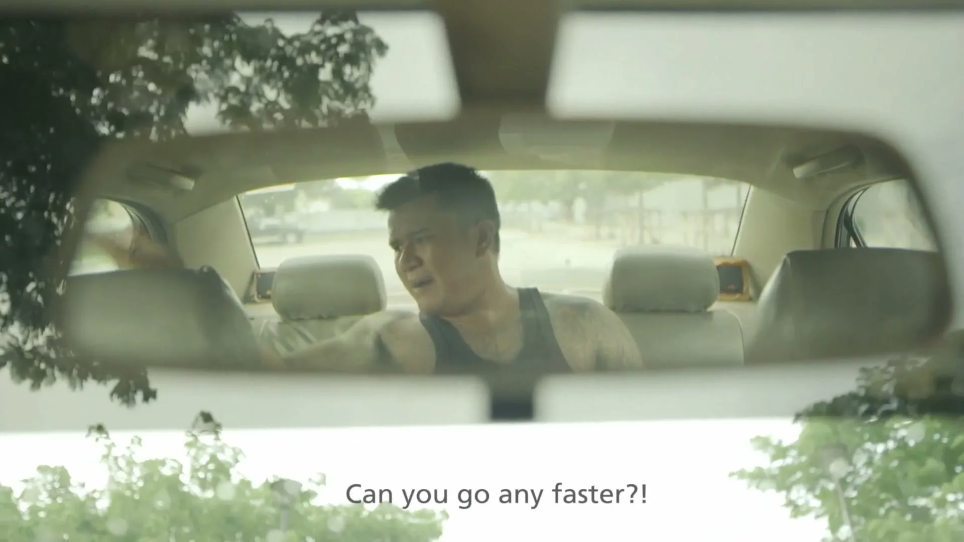 A male passenger in a tank top, seen through a car's rearview mirror, looks agitated and gestures in the back seat of a taxi.