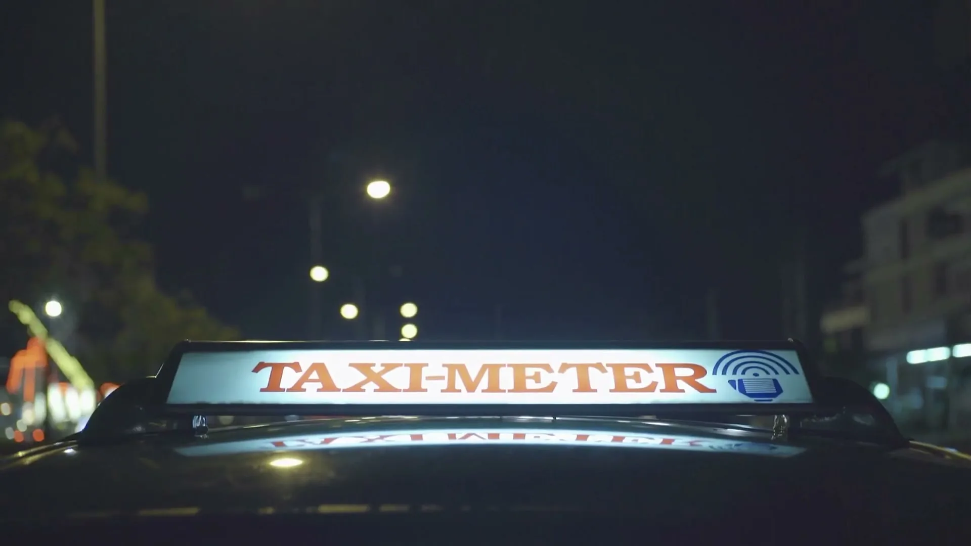 A close-up shot of a "TAXI-METER" sign on top of a taxi at night, with blurred city lights in the background.