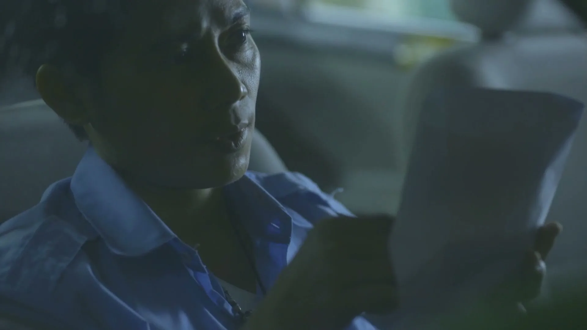A middle-aged person in a blue shirt sits in a car at night, looking at a piece of paper with a thoughtful expression.