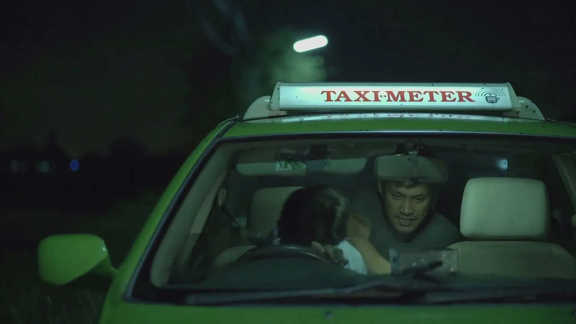 A young boy sleeps on his father's lap in the front seat of a green taxi at night, while the father looks at his phone.