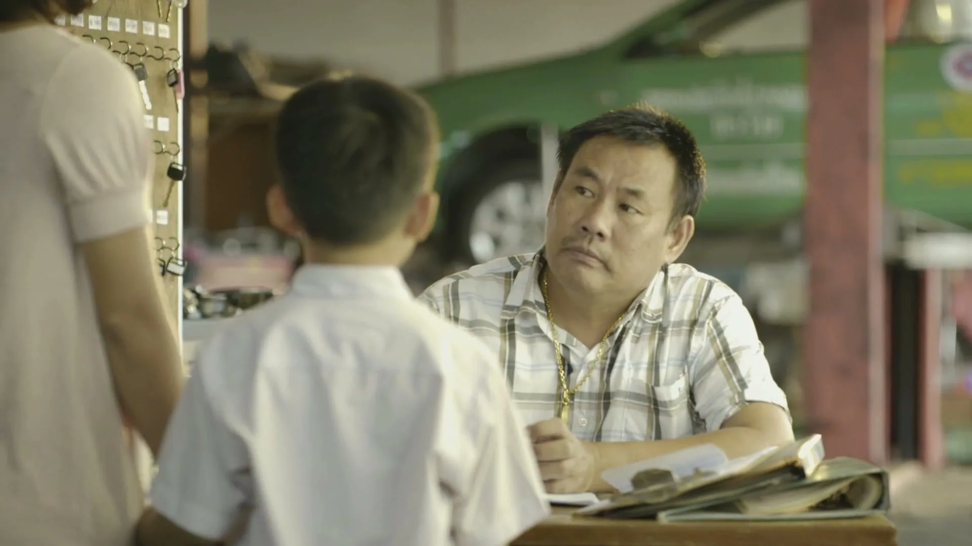 A man in a plaid shirt sits at a desk, looking up at a young boy in a white shirt, while a woman stands nearby in a garage.