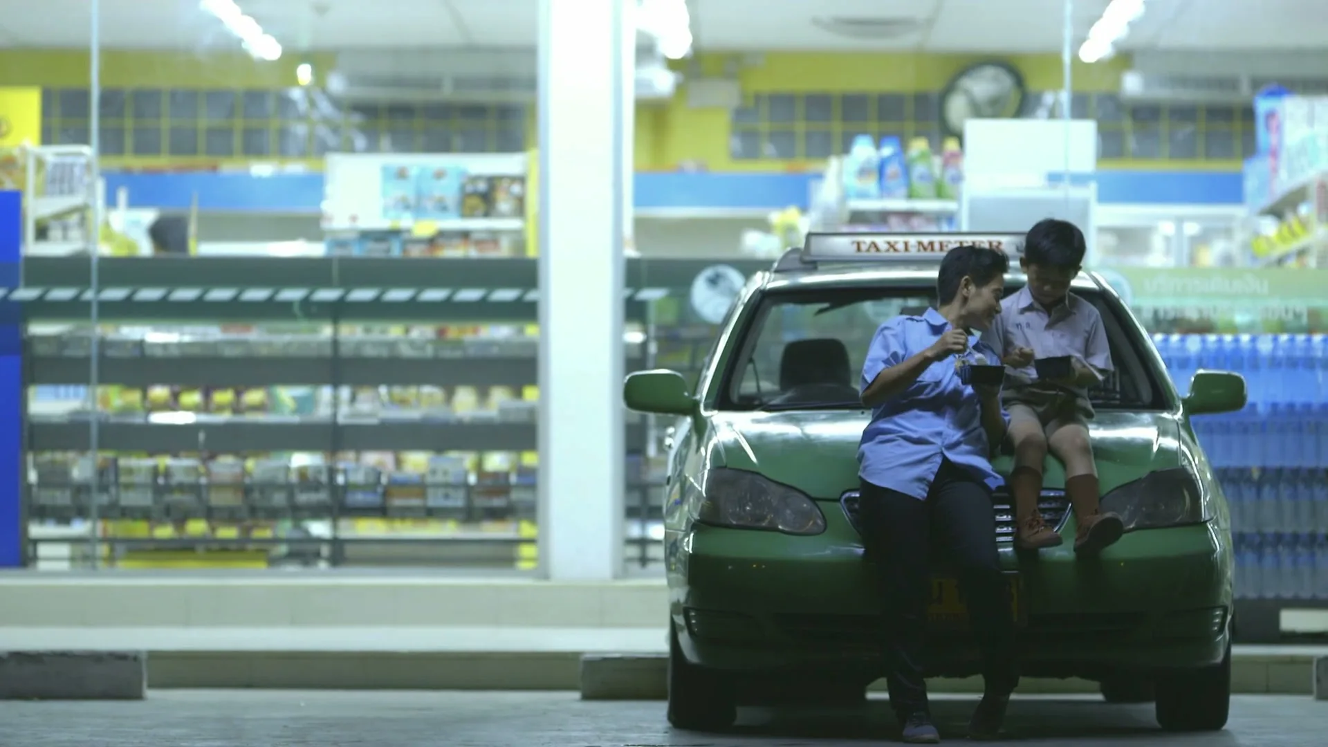 A taxi driver and a young boy sit on the hood of a green taxi, both looking at their phones in front of a convenience store at night.