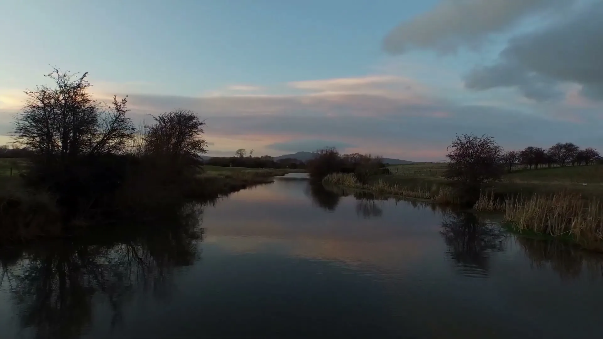 A wide shot of a calm river reflecting the colorful sky at dusk, surrounded by bare trees and grassy fields.