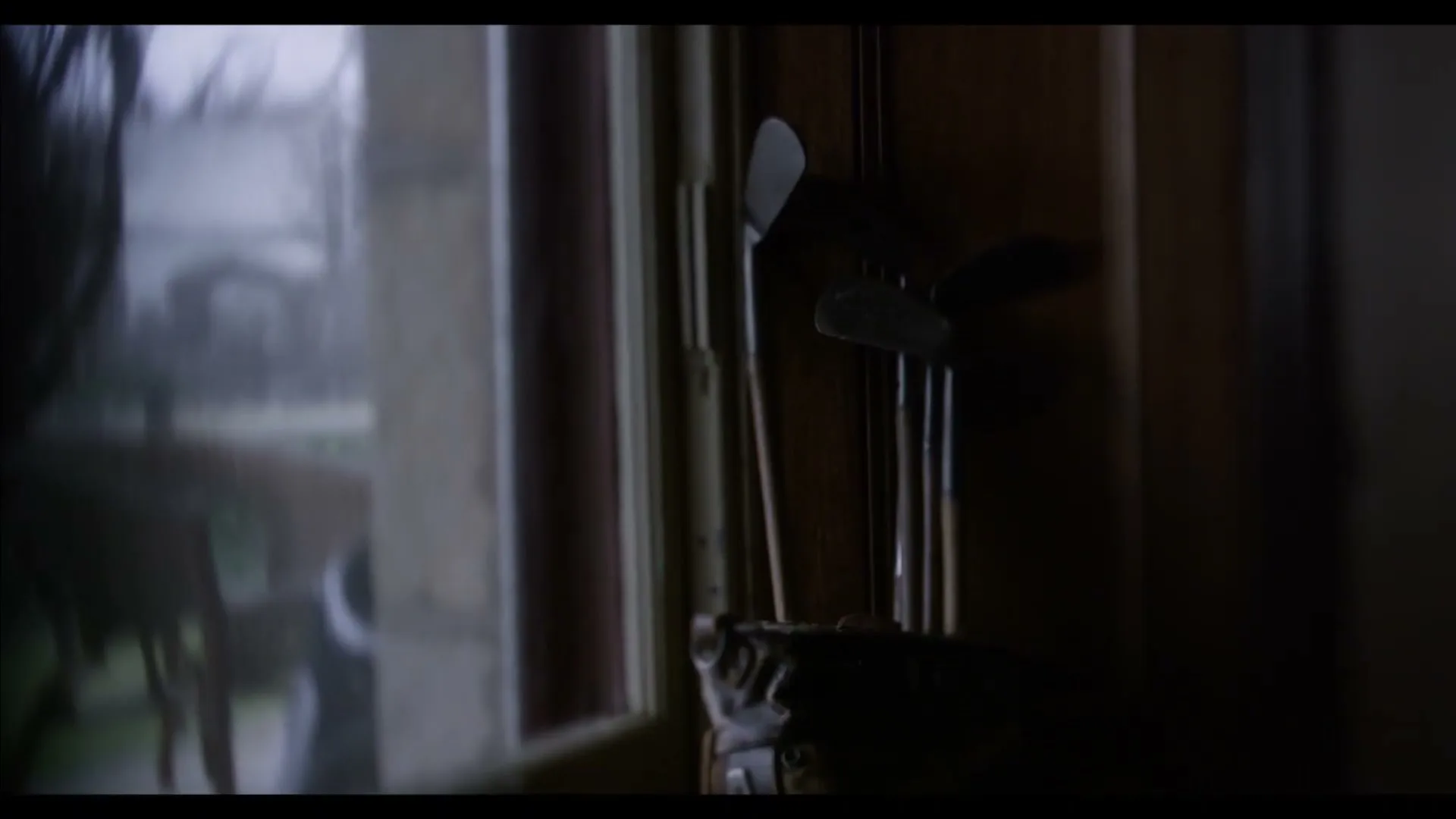 A set of golf clubs stands in a dark corner next to a window, with a blurry outdoor scene visible through the glass.