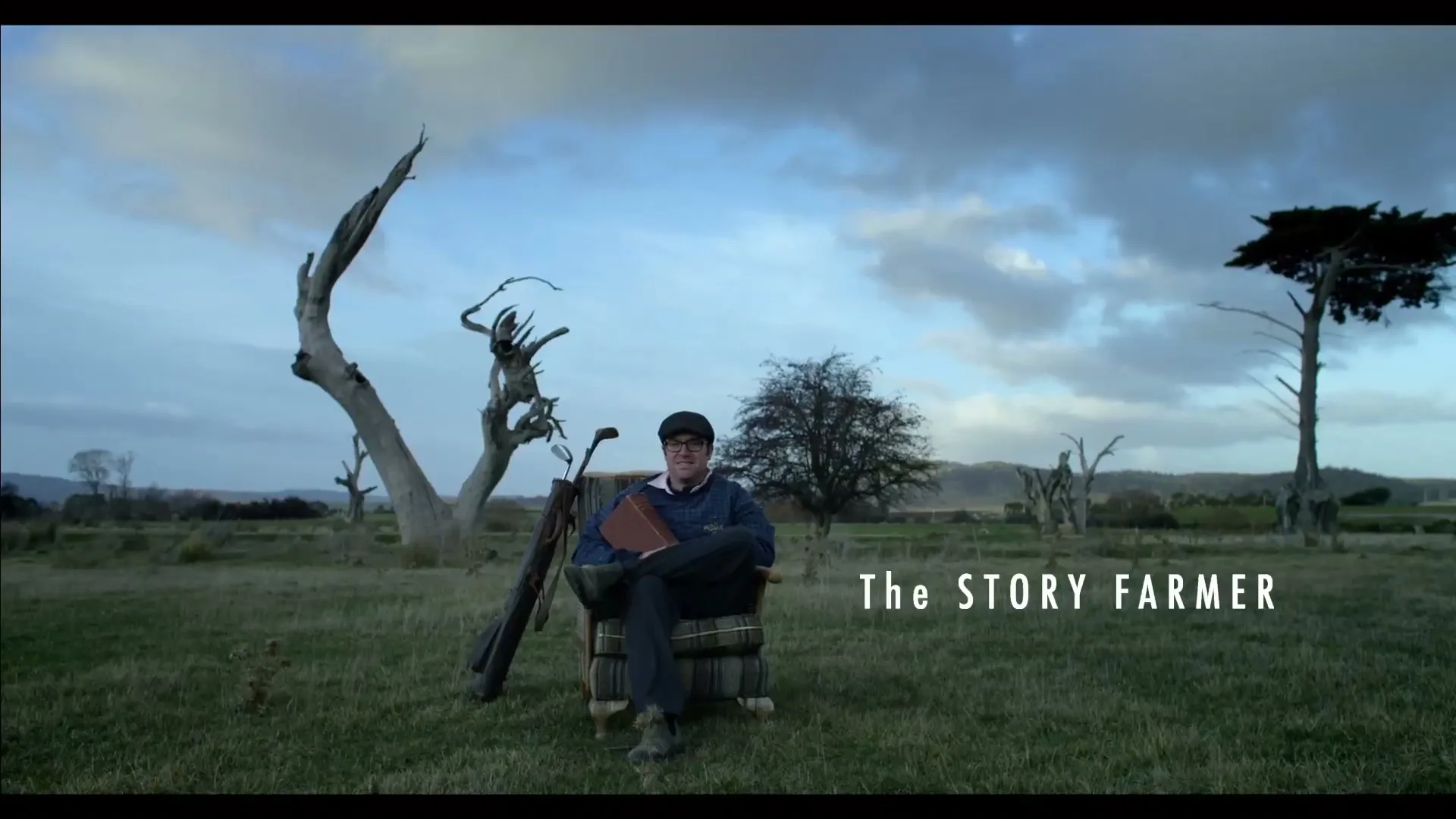 A man in a blue sweater and cap sits in an armchair in a grassy field with dead trees and a golf bag next to him, under a cloudy sky.