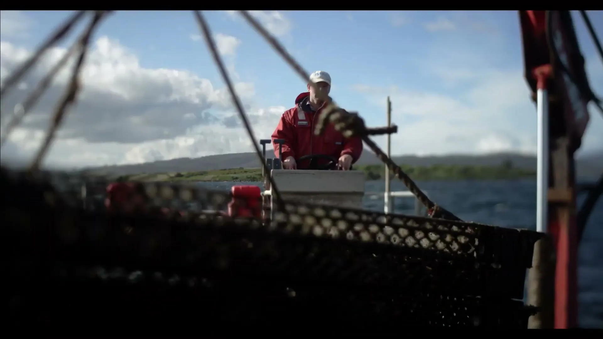 A male oyster farmer in a red jacket and white cap steers a boat on a sunny day, with a blurred foreground of oyster cages.