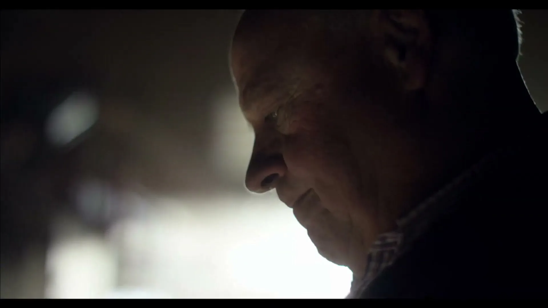 A man in his 60s is seen in profile, looking down with a gentle smile, in a dimly lit interior setting.