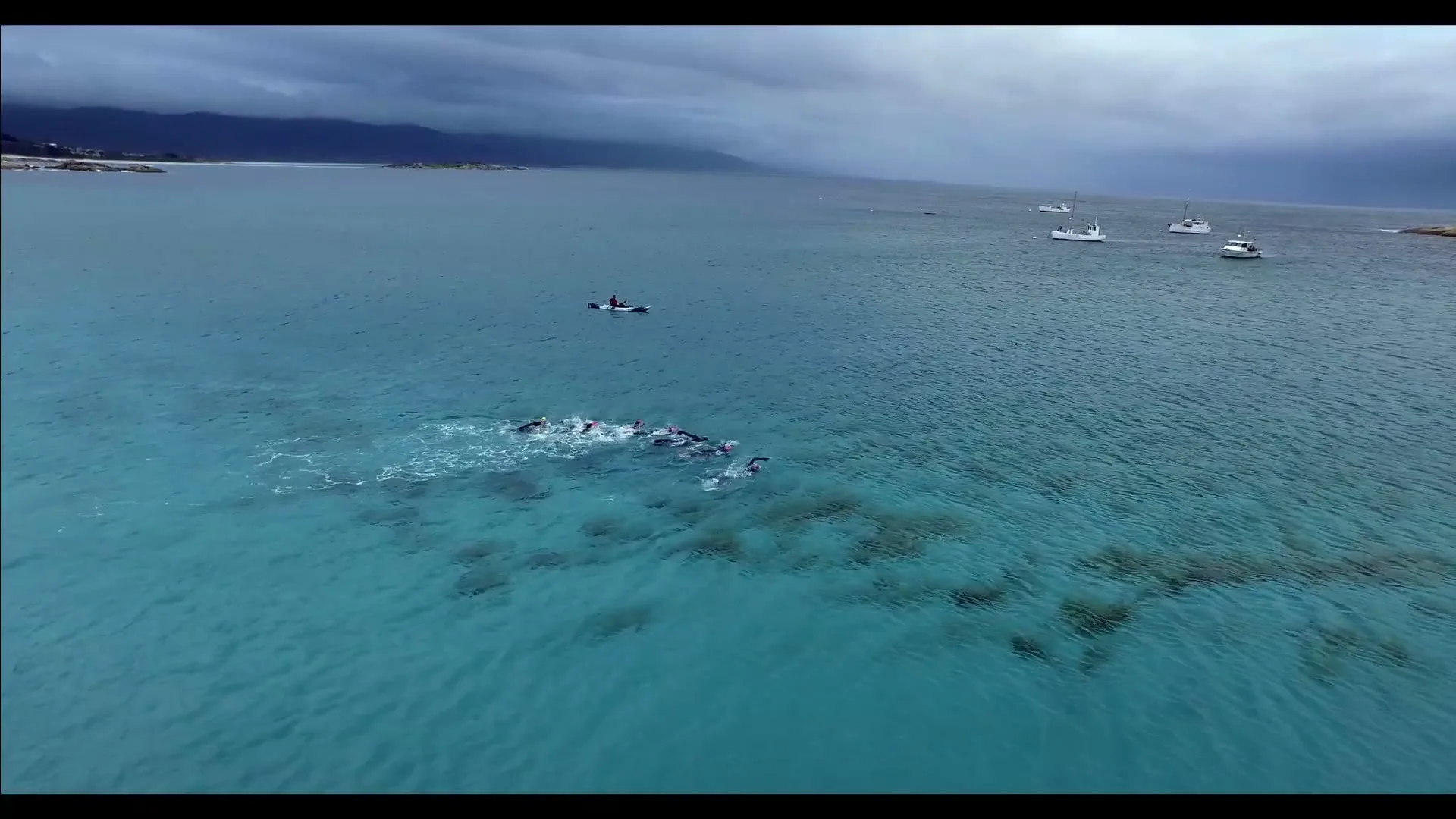 An aerial view shows a group of swimmers in wetsuits swimming in clear blue ocean water, with a small boat and mountains in the background under a cloudy sky.