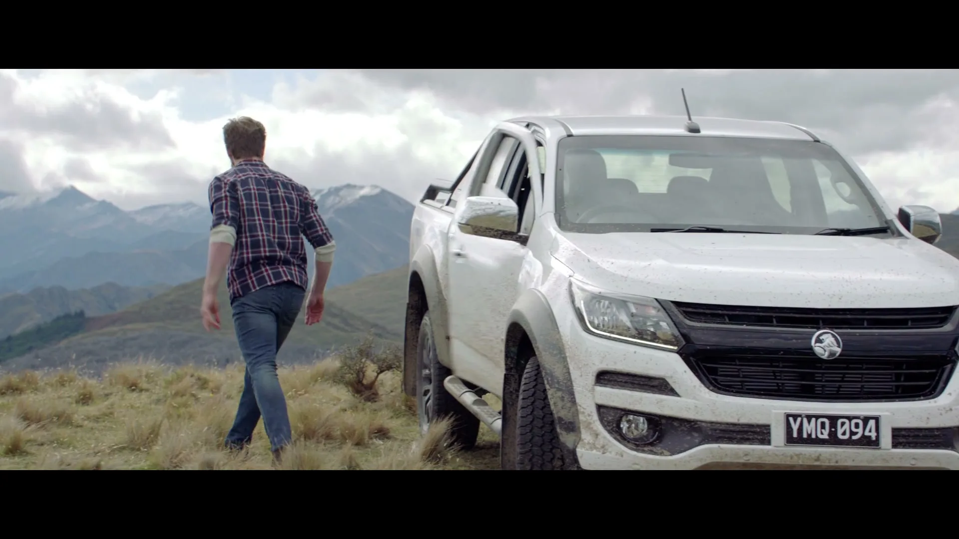 A man in a plaid shirt and jeans exits a white pickup truck on a grassy mountain peak, looking out at the scenic landscape.