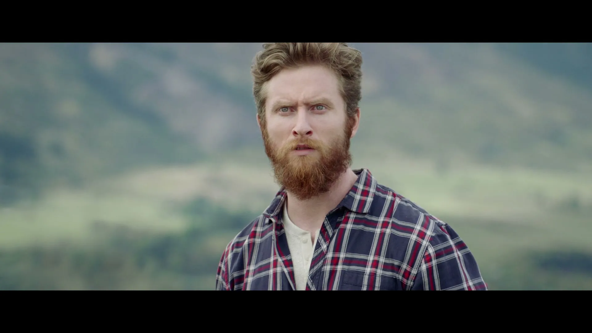 A man with a red beard and a plaid shirt looks confused in a wide outdoor shot with mountains in the background.