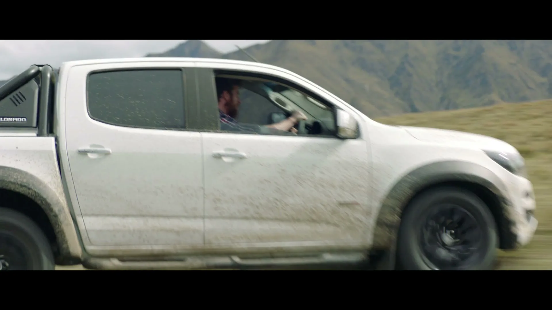 A man with a beard drives a white pickup truck up a grassy hill, with mountains in the background under a cloudy sky.