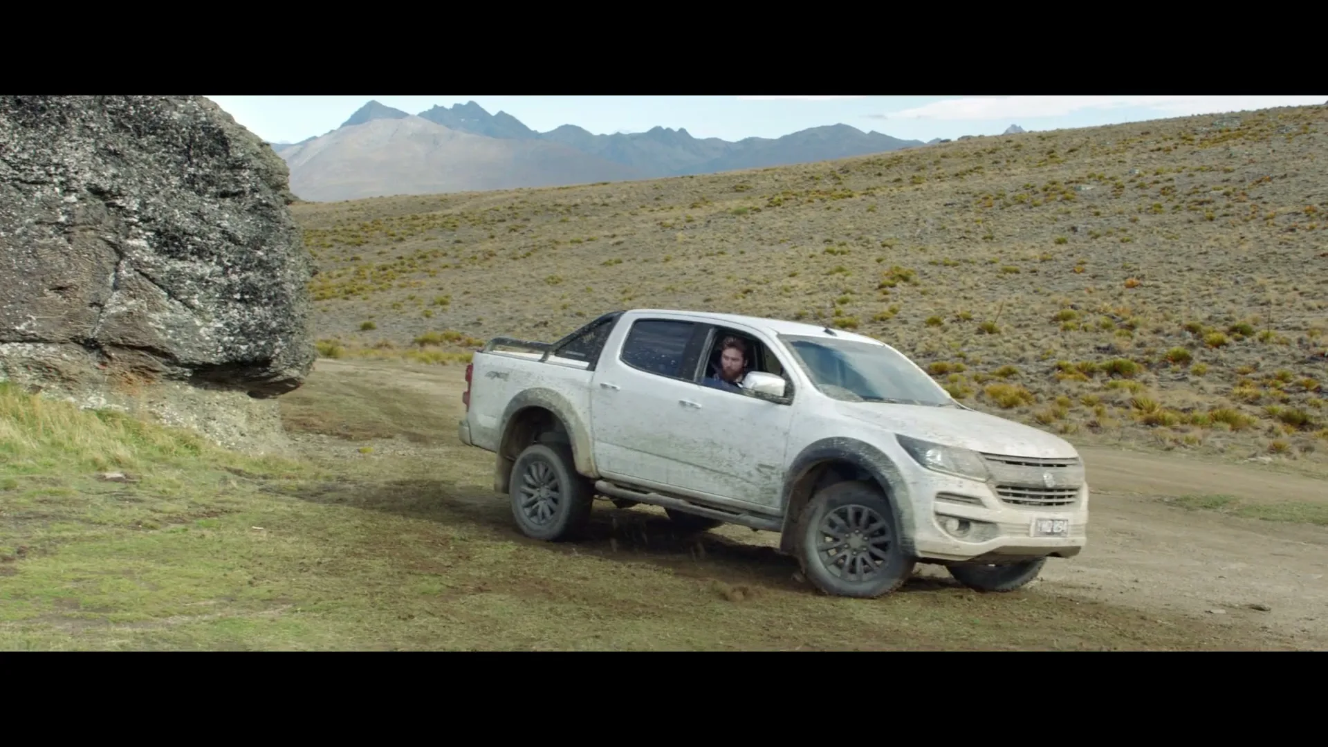 A white Holden Colorado pickup truck drives slowly up a dirt path next to a large rock, with mountains in the background.