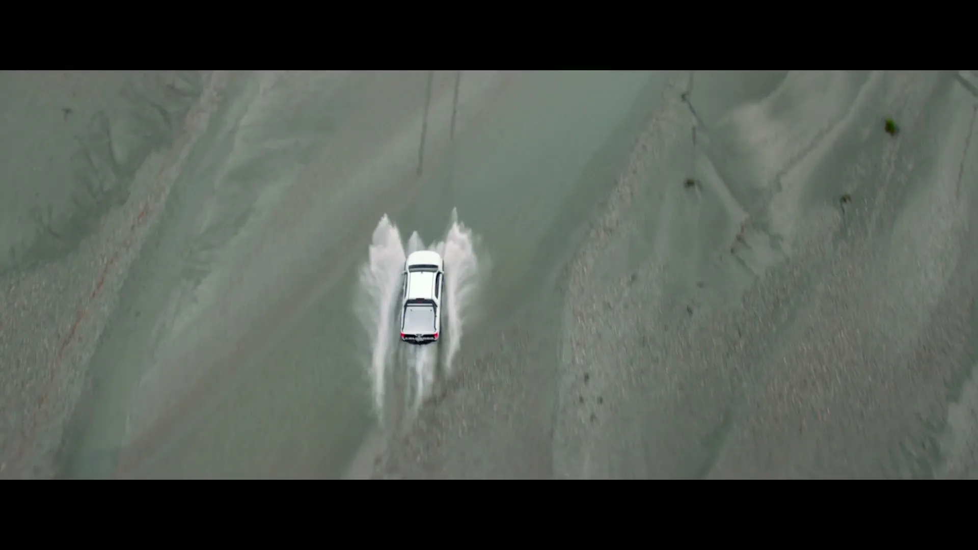 A white Holden ute drives through a shallow river, creating splashes of water, in an extreme wide overhead shot.