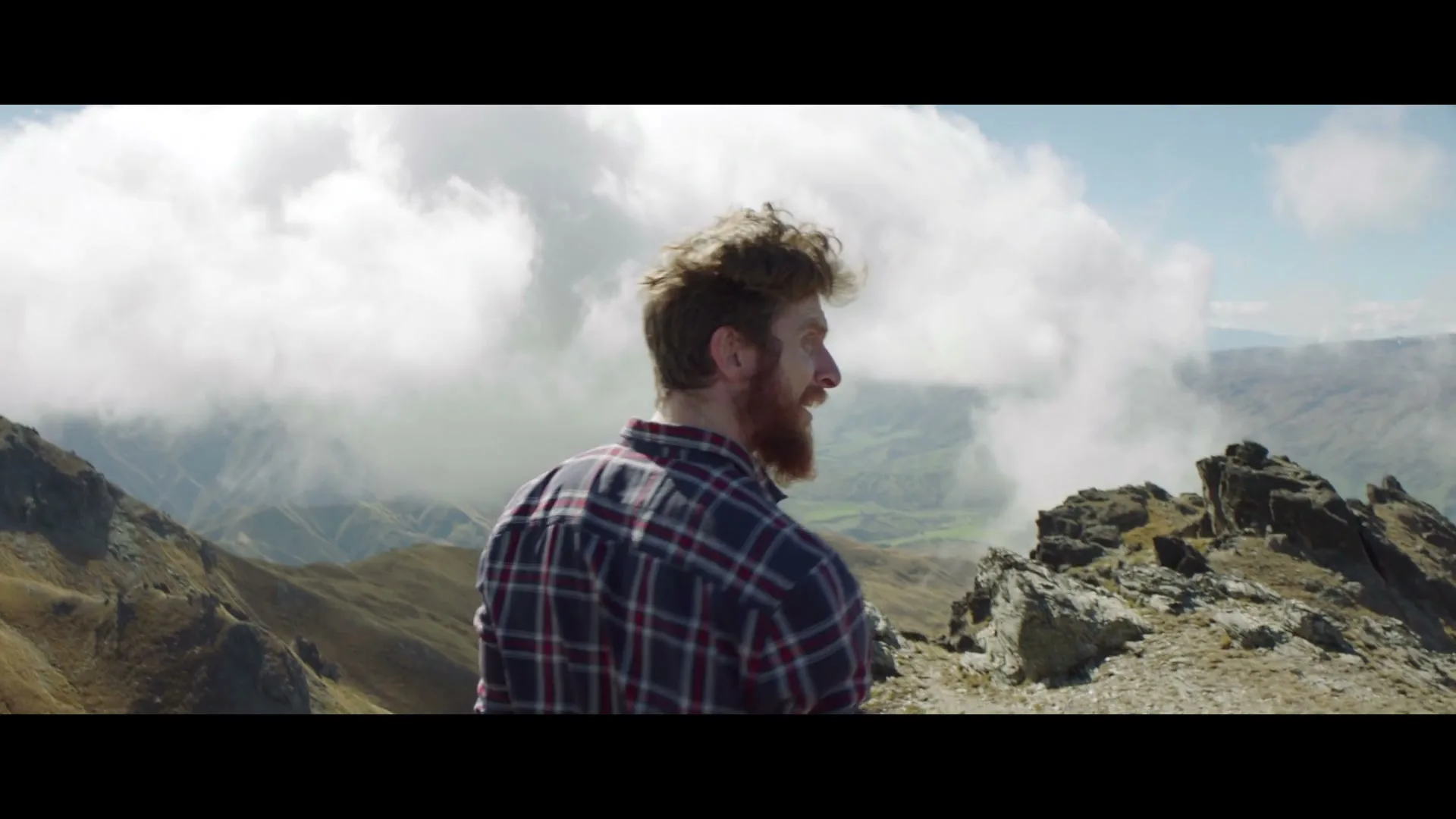 A man with a beard and plaid shirt stands on a mountain peak, looking out at a cloudy, mountainous landscape on a sunny day.