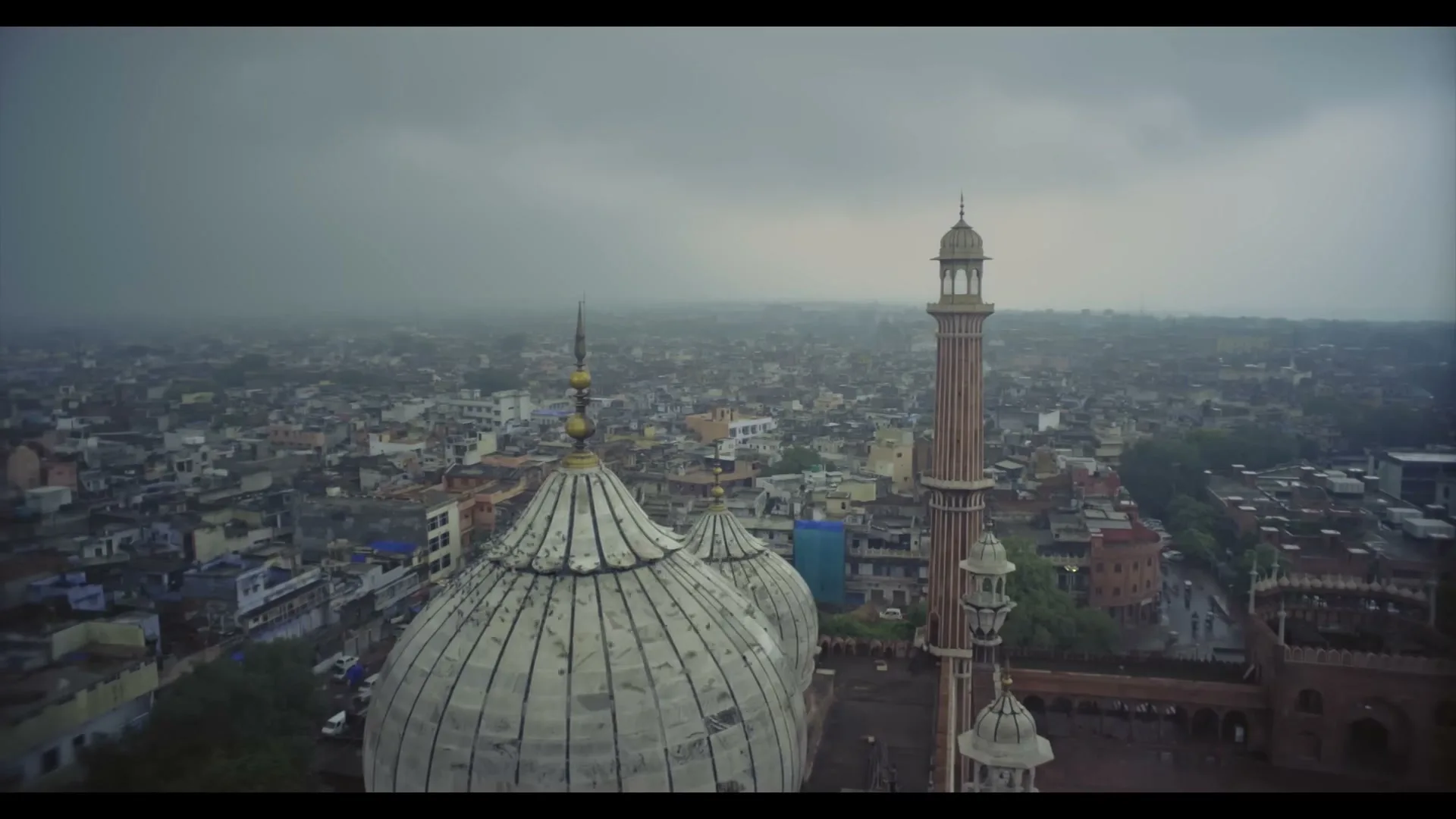 An aerial view of a large mosque with a white dome and a tall minaret, surrounded by a dense city under a cloudy sky.