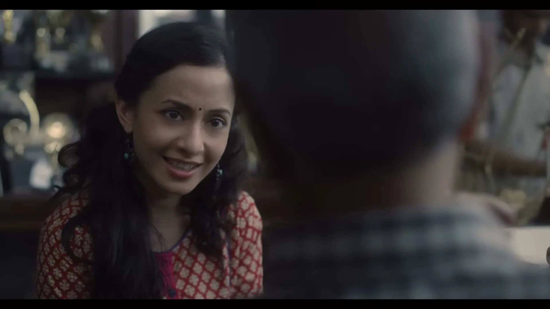 A young Indian woman with long dark hair and a red patterned top smiles and talks to an older man, whose back is to the camera, in a shop.