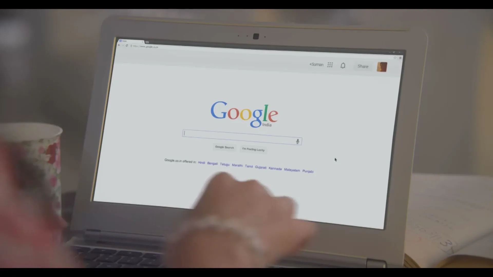 A person's hand types on a white Samsung laptop, displaying the Google India search page, on a desk with a floral mug.