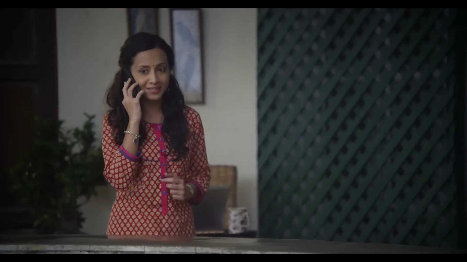 A woman in her 30s, wearing a red patterned tunic, talks on a smartphone with a concerned expression in an interior setting.
