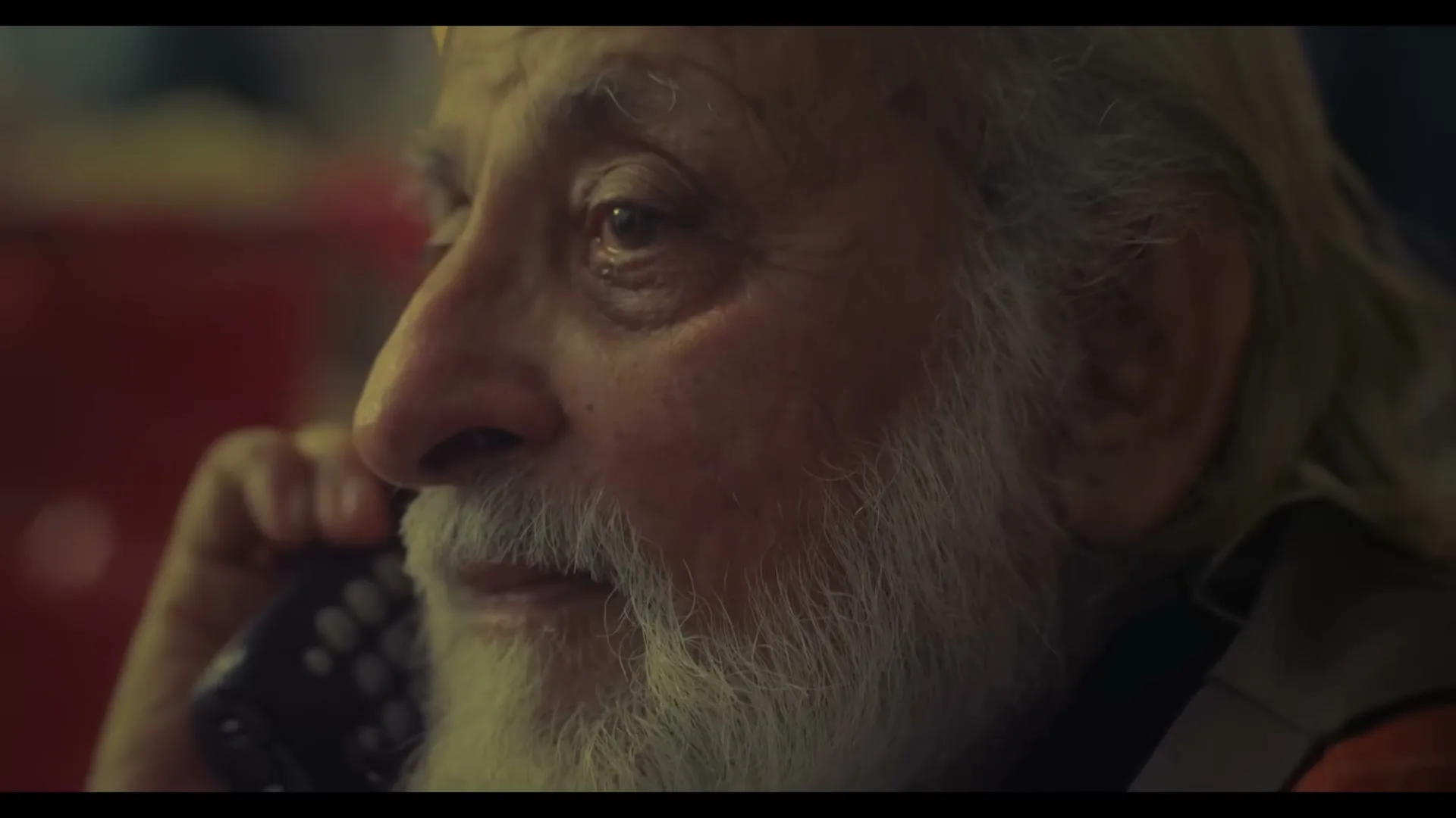 An elderly man with a long white beard and hair holds a phone to his ear, listening intently with a slight smile.