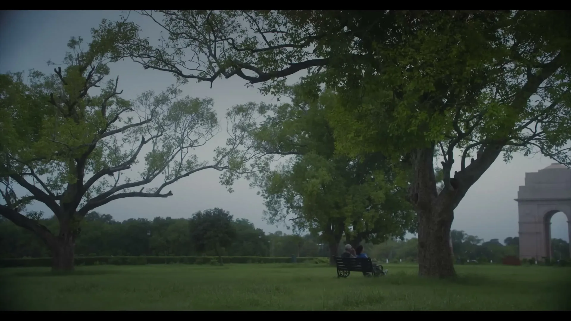 Two elderly men sit on a park bench under large trees, with a distant monument visible on the right side of the frame.