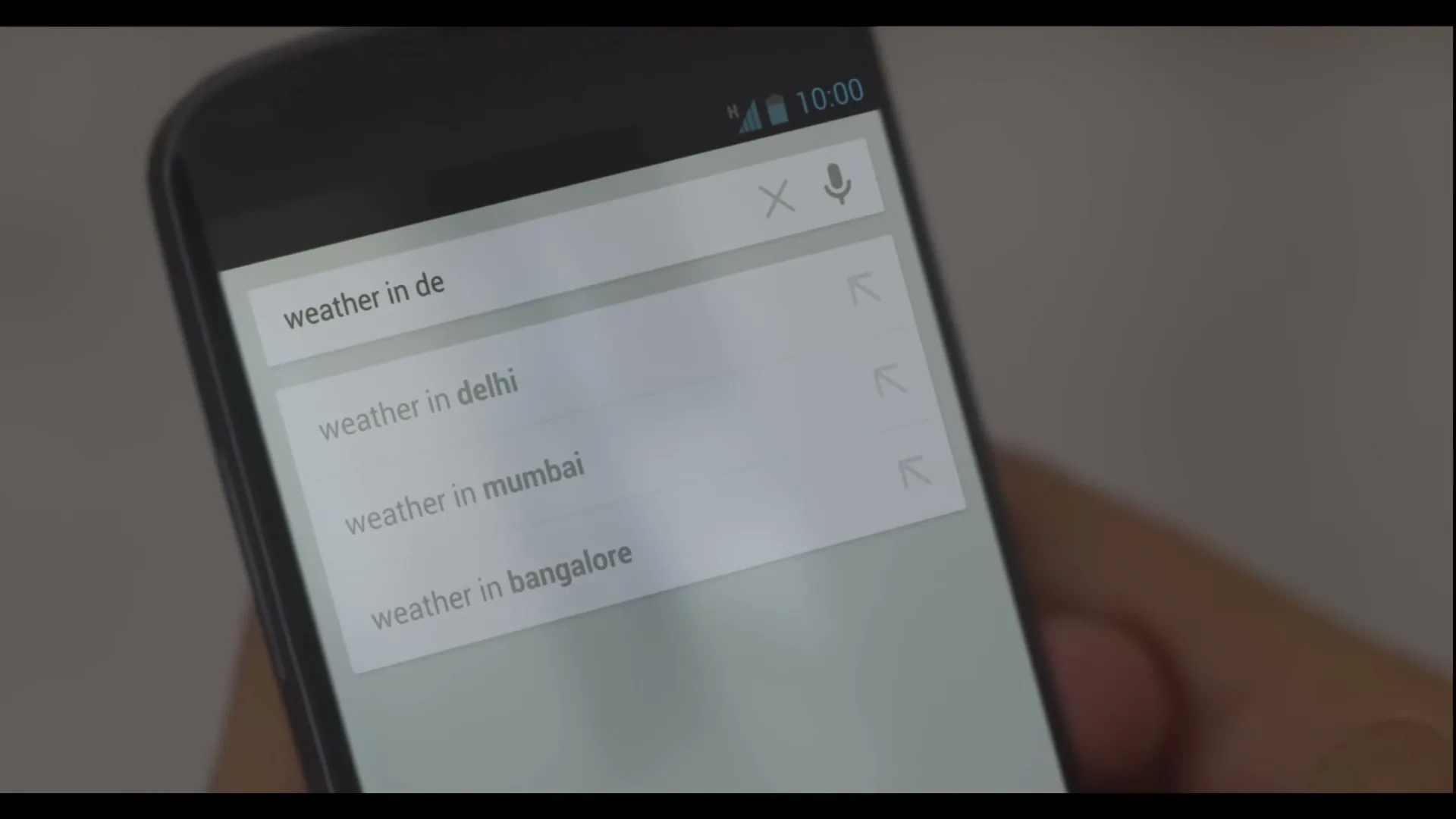 A person's hand holds a smartphone, typing "weather in delhi" into the Google search bar, revealing weather information for Delhi, India.