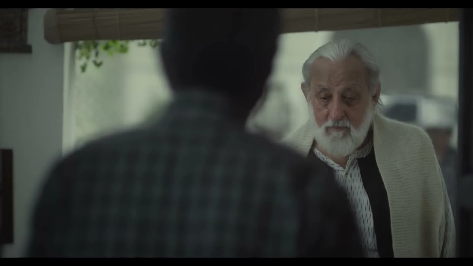 An older man with a white beard and a shawl looks at a younger man with a concerned expression in a home.