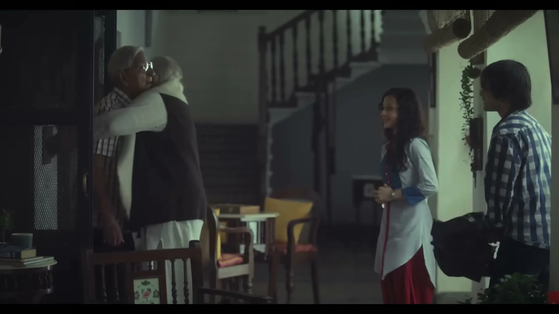 Two elderly men embrace warmly in a dimly lit home, while a young woman and a young man watch them with smiles.