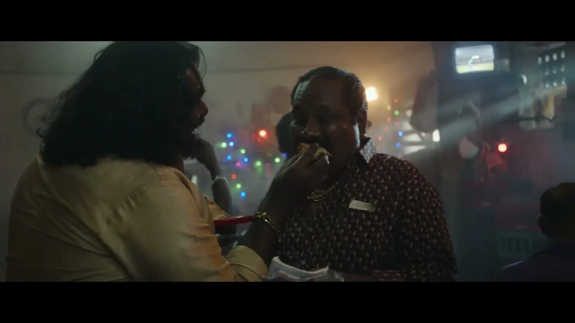 Two men are seen in a dimly lit, smoky room with colorful lights, one feeding the other a piece of food while holding money.