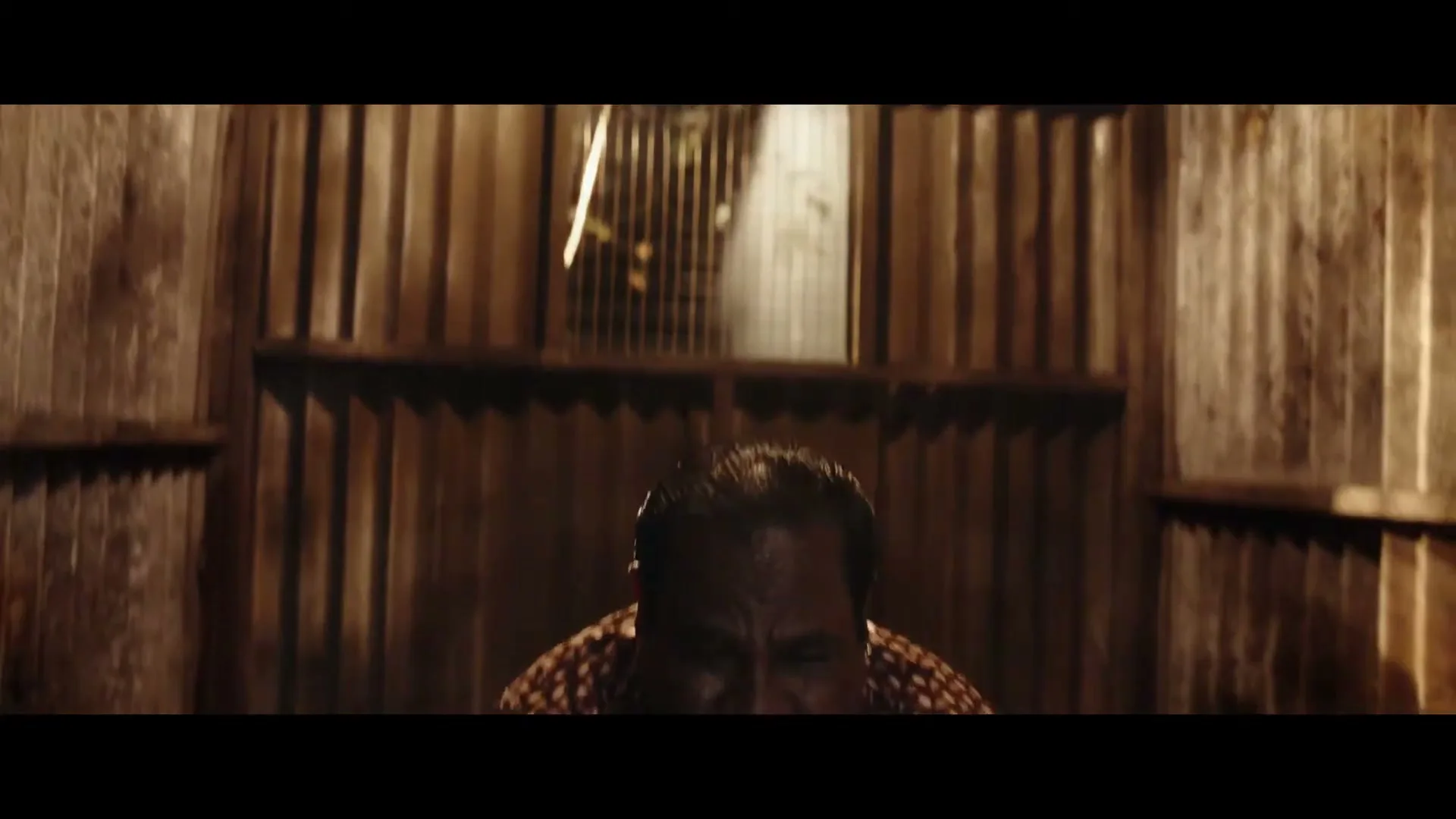 A man in his 50s, wearing a patterned shirt and gold chains, looks up with a surprised expression in a dark, corrugated metal room.