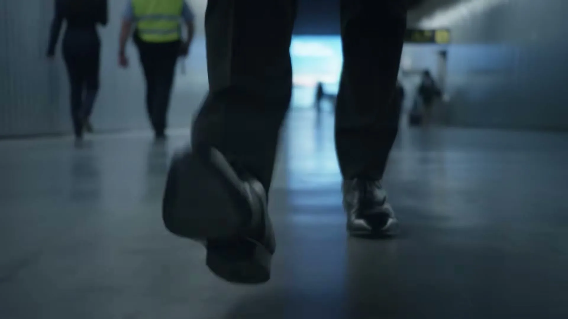 A low-angle shot follows a person's legs and feet as they walk down a dimly lit hallway, with other blurry figures in the background.