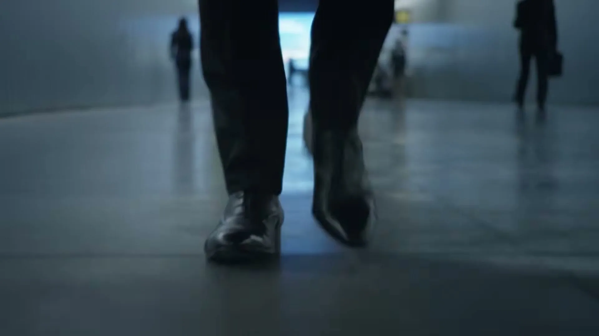 A low-angle shot shows a person's feet in dress shoes walking down a dimly lit hallway, with other blurred figures in the background.