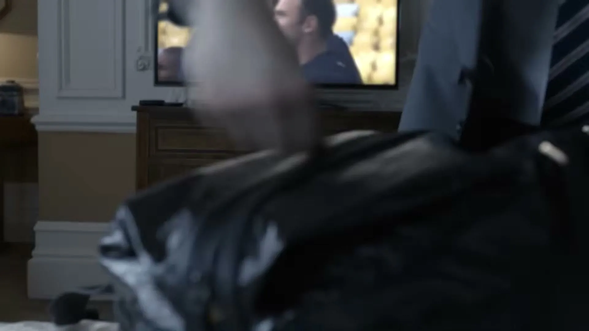 A man in a suit and tie packs a black bag while a soccer player is visible on a TV screen in the background.