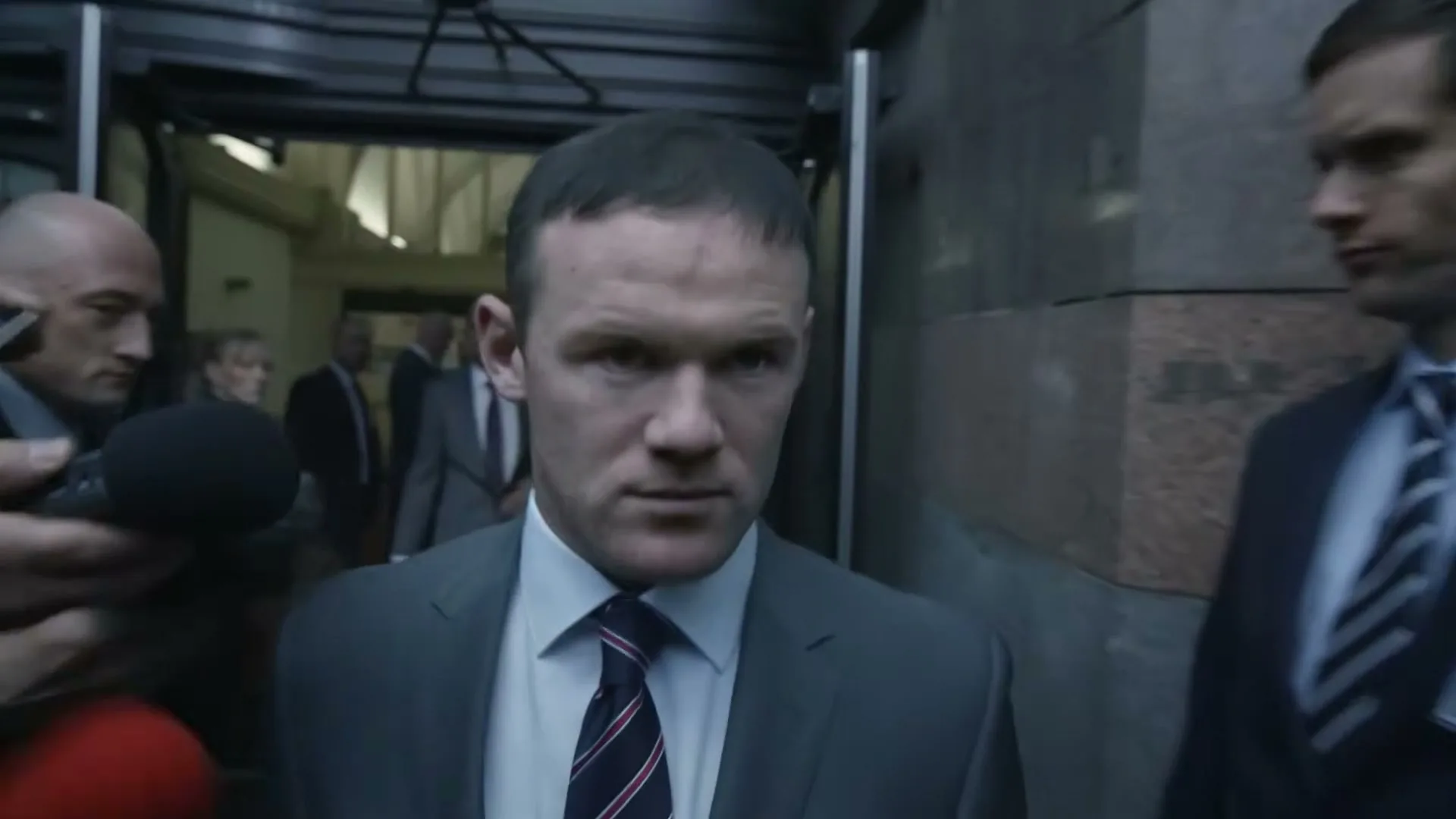 Wayne Rooney, a male in his 20s, wearing a suit and tie, walks through a crowd of paparazzi and reporters outside a building.