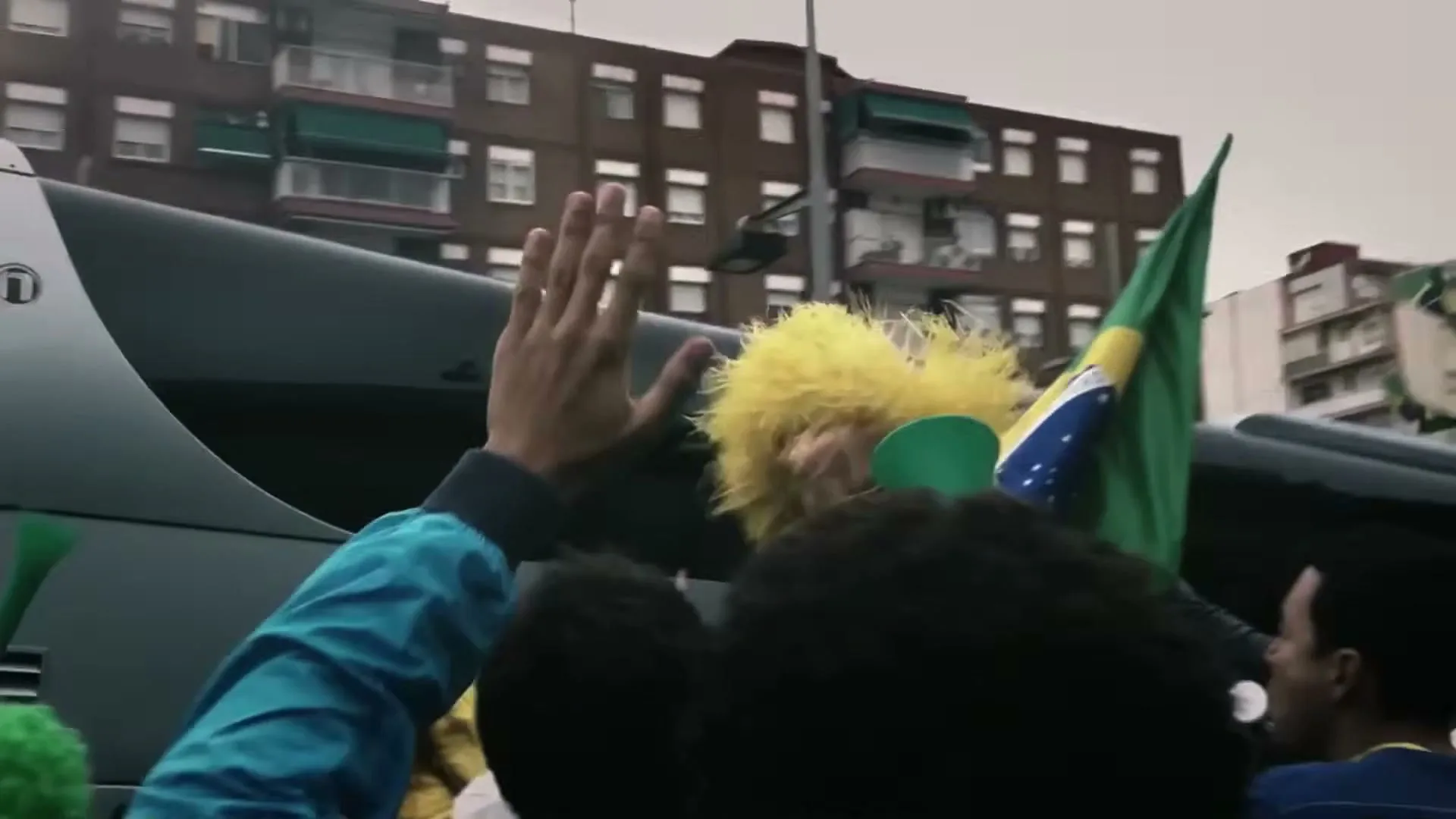 A crowd of Brazilian fans, some wearing yellow wigs and waving flags, cheer from a moving bus on a city street.
